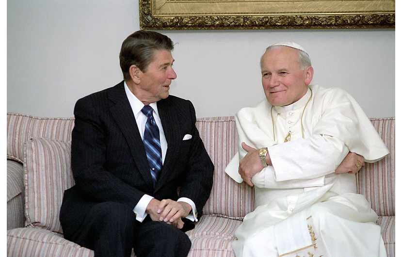 An in-color photograph of U.S. President Ronald Reagan Meeting with Pope John Paul II at The Fairbanks Airport in Alaska in 1984; both sit on a pink colored sofa, at the left side of the picture sits Reagan looking directly at the pope; on the right side sits J.P. II with his arms crossed looking ahead.
