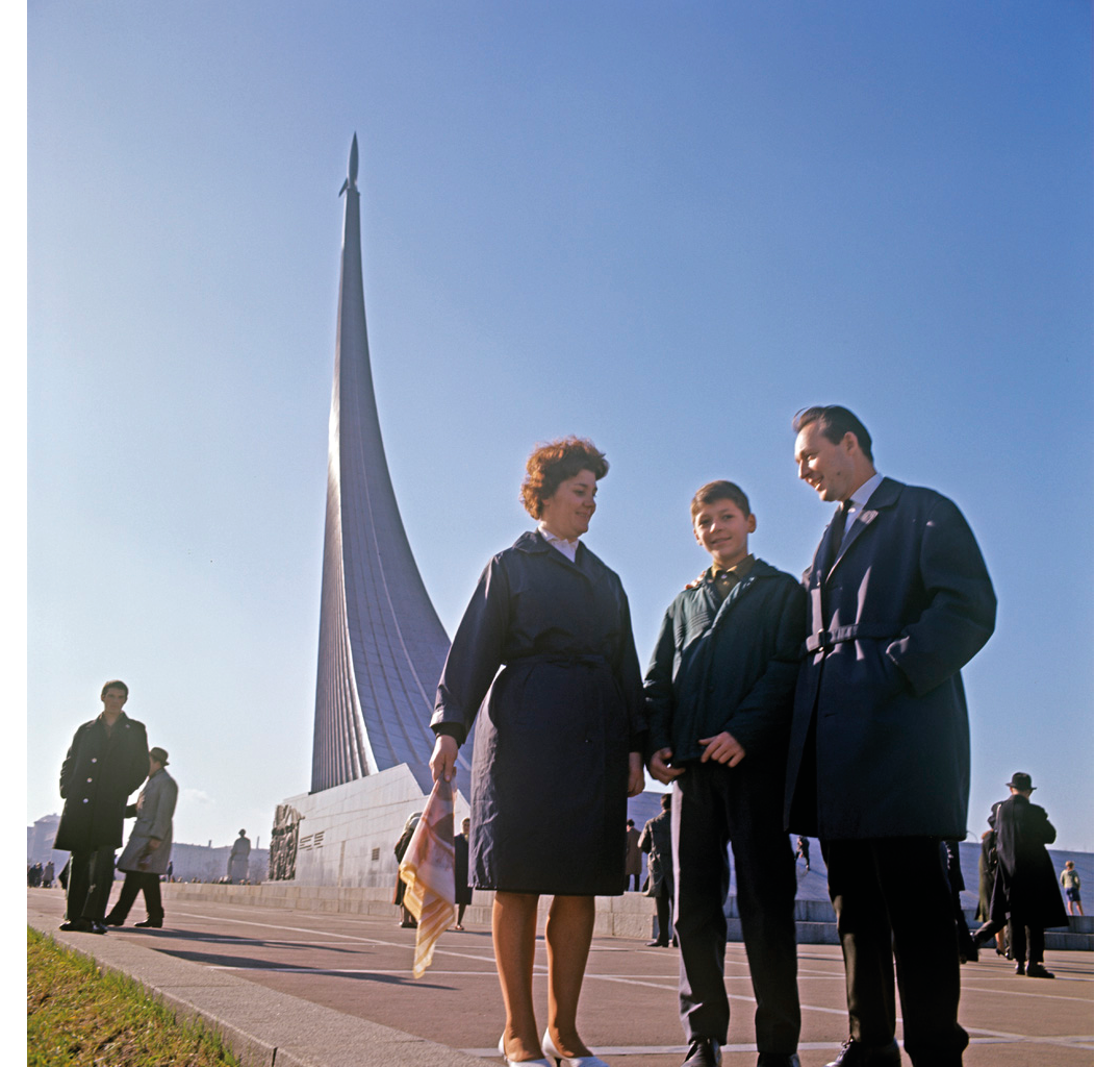 An in-color photograph from 1964: family poses in front of the Monument to the Conquerors of Space in Moscow; the man, with his hands in his trench coat pockets, looks at the woman holding a headscarf in her right arm while looking at the young boy who looks directly into the camera; background shows other monument visitors and clear blue skies.