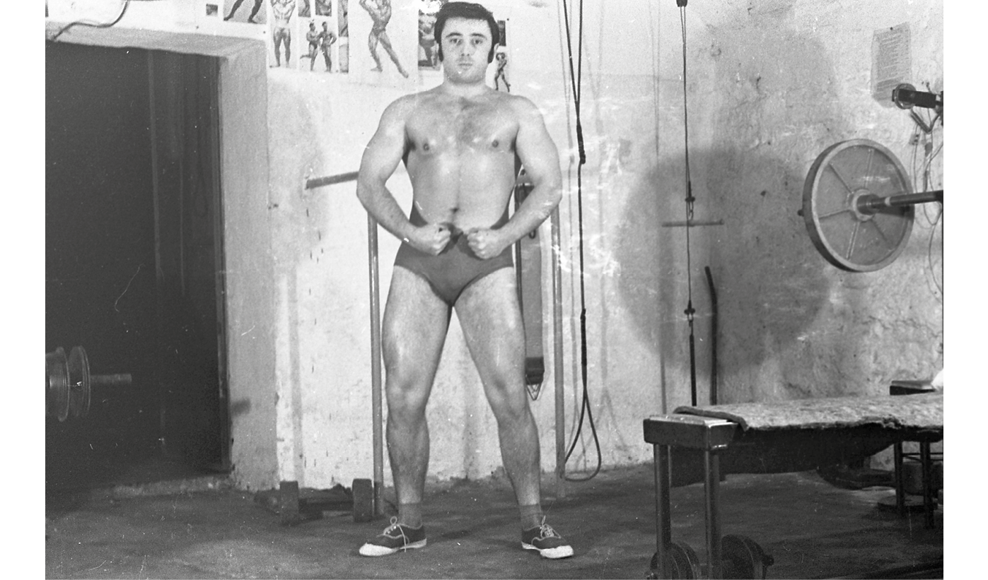 A black and white photograph from 1970 Budapest of bodybuilder Tamás Urbán inside the Gépgyár Sport Klub gym: shows a shirtless Urban flexing his arms and legs for the camera surrounded by gym equipment.
