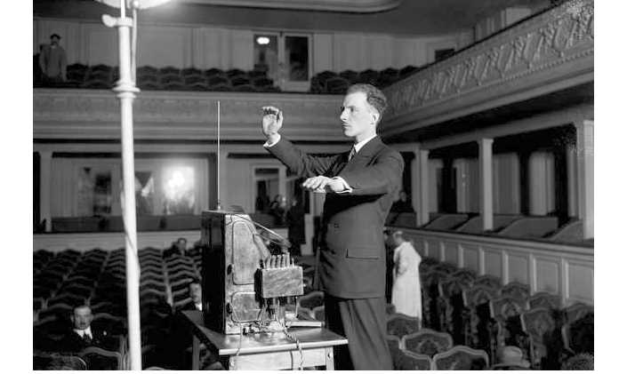 A black and white photograph; shows inventor Leon Theremin in a black suit, demonstrating his instrument; he stands in the middle of a venue with a small scattered audience and many empty chairs.