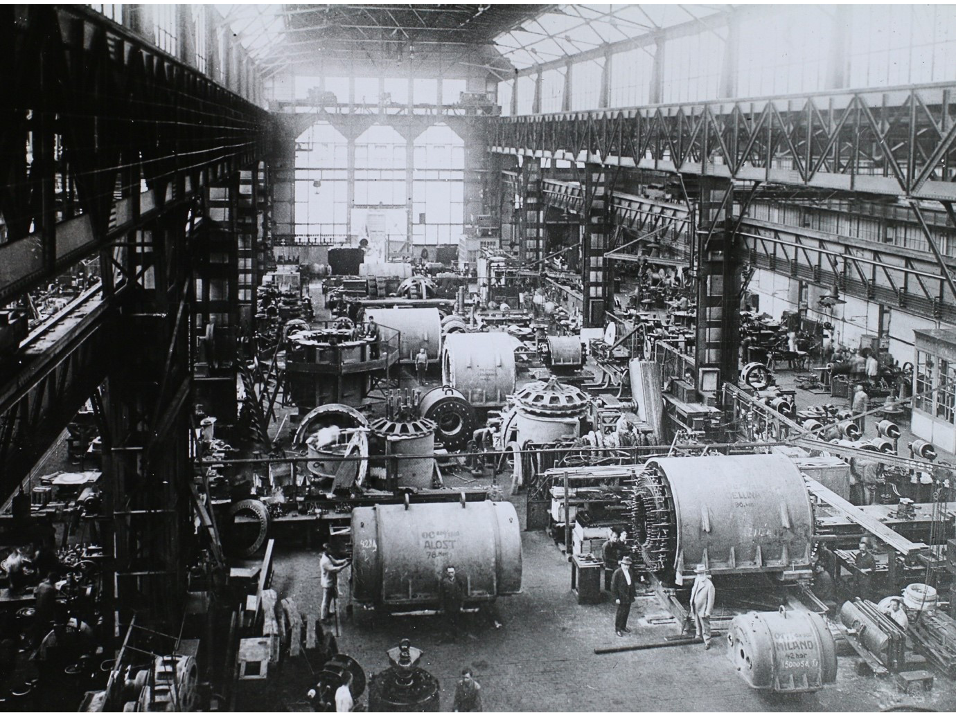A black and white picture of the Ganz factory hall in 1922 Budapest; shows hall with iron rails on both sides with various types of industrial machinery managed by a few workers at the center; three large windows at the back and smaller windows around the roof illuminate the hall.