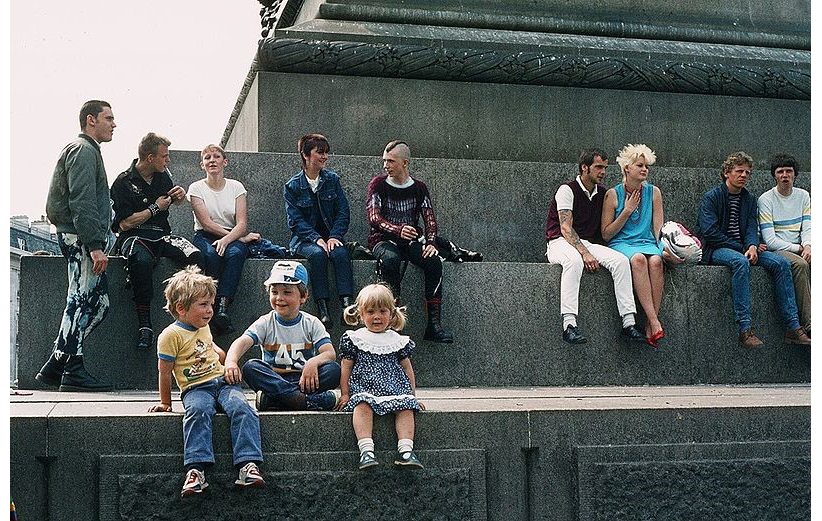 A photograph in color from 1983 London: shows people standing and sitting at the base of Nelson's Column in Trafalgar Square; three young children sit at the first step of the monument; 8 older people sit at the second step while one at the far side left stands.