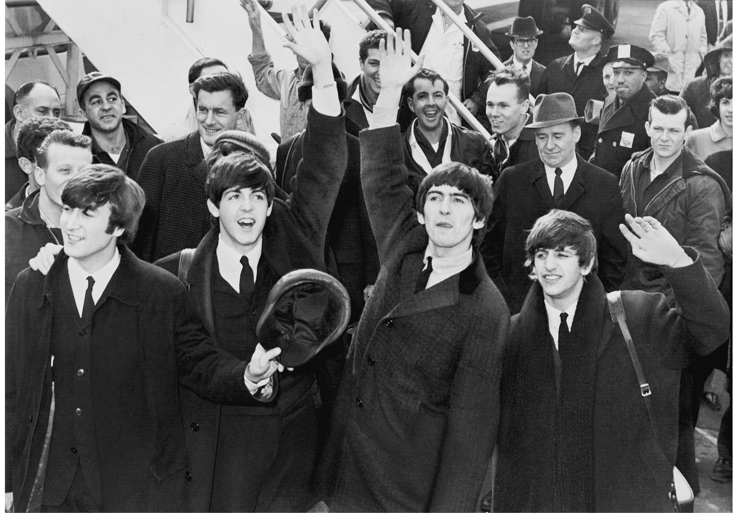 A black and white photograph from 1964 of The Beatles after arriving at Kennedy Airport shows the four music group members waving to their fans; behind them, other flight passengers walk from the airstair.
