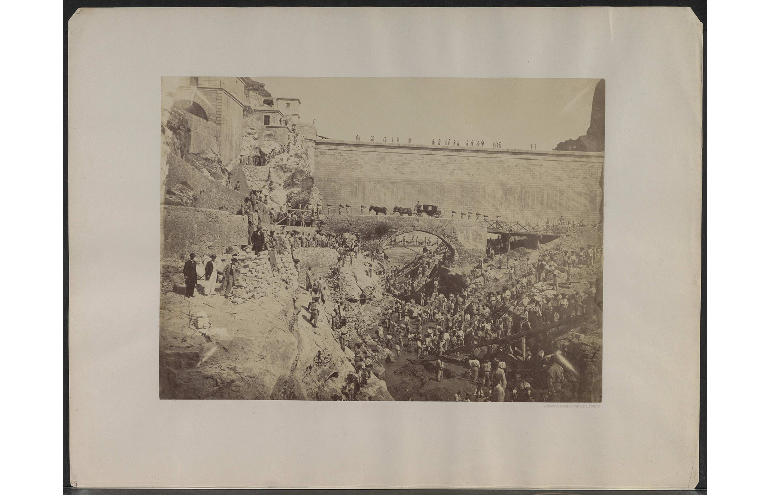 1850s black and white photograph: shows a dam worksite in Madrid; on the left side, men in suits pose for the camera; rest of the image depicts a line of workers as they walk from the top of the dam all the way down to the canal; in the back, a carriage is drawn by two mules/horses across a bridge connects both sides of the canal.