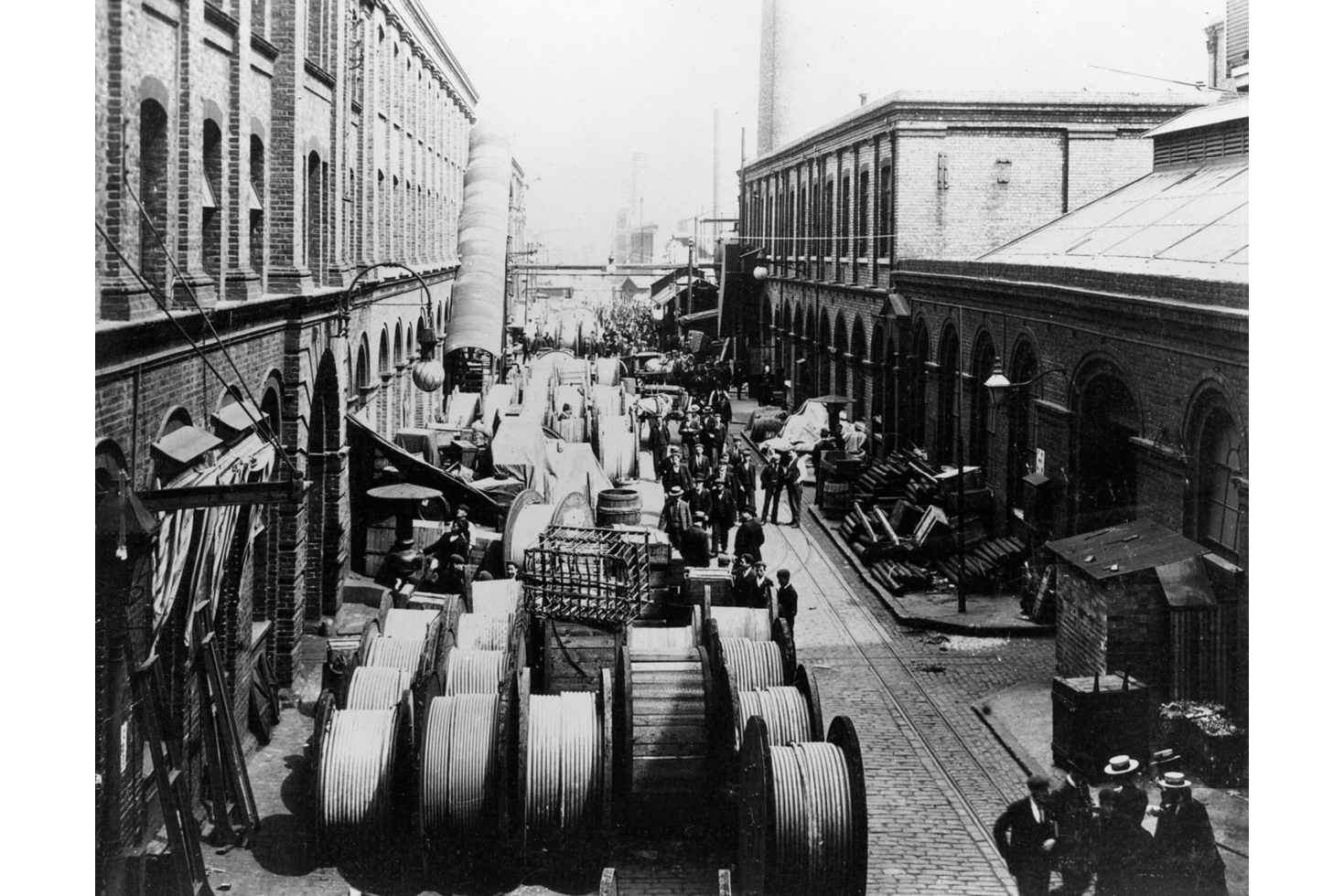 A black and white photograph of Siemens Brothers Ltd., Woolwich Works near London ca 1890: shows a cobbled street outside of an industrial building filled with wrapped cable lines; various men in suits make their way through the streets as they chat and observe the lines.