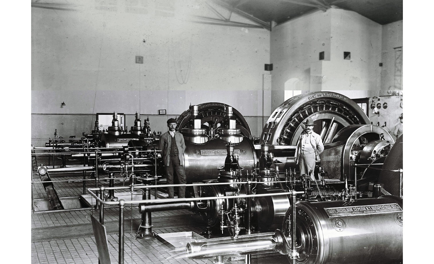 A black and white photograph from 1914 Hungary: shows a steam engine factory with two men posing for the photograph next to one of the machines.