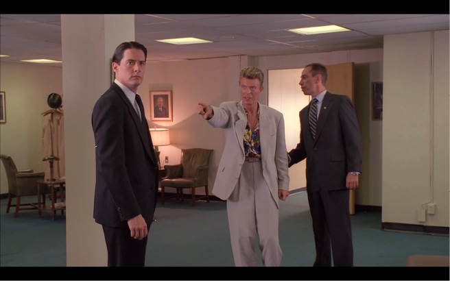An image of what appears to be an office space, three men stand in the centre. The first man, left, stares past the viewer with a shocked expression on his face. The man in the centre points at the first man as if accusing him of something. The third man, right, leans behind the man in the centre, holding the arm not pointed at the first man - he stares off to the left of the image, at something out-of-shot.