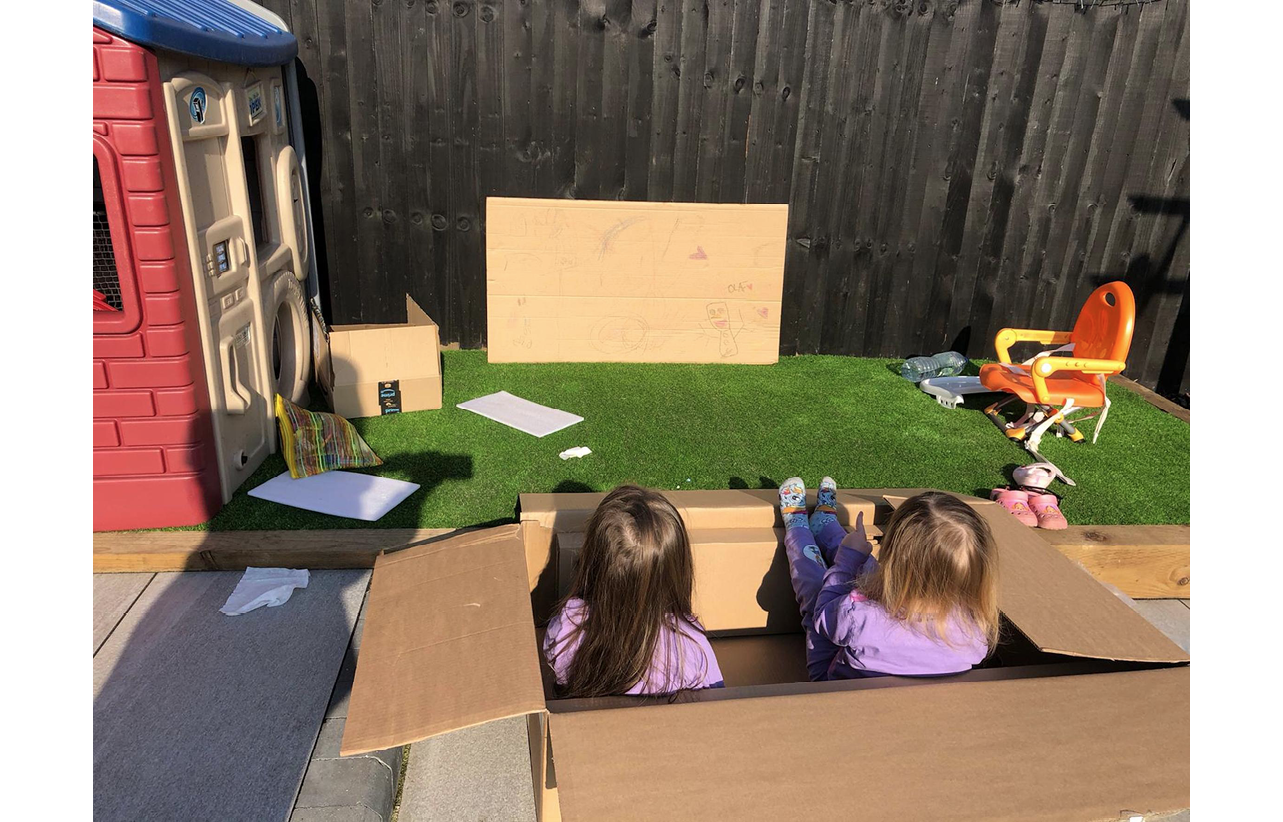 Photo of two children in a garden, sitting in a large cardboard box, watching a pretend cinema screen made of cardboard, propped against fence
