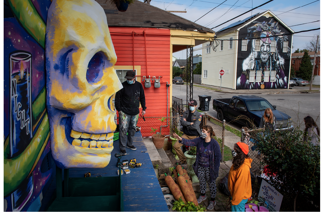 The photo is taken from one end of a porch, looking along the front of a house on Toledano Street, New Orleans. Four people wearing paint-splattered clothes and face coverings are standing on or in front of the porch, facing the in-progress house float. Float designer and cofounder of the Hire A Mardi Gras Artist initiative Caroline Thomas is among them, with her right arm outstretched. A large paper-mâché skull in relief, painted in yellow and purple, hangs on the front of the house. A small sign waiting to be hung reads ‘House #1, The Night Tripper’, the name of an album by late New Orleans musician Dr John. A mural of Dr John is painted on a house across the street in the background.