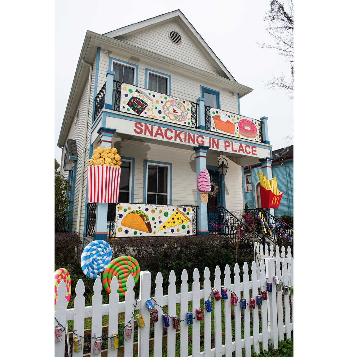 A two-storey white house with blue trim, a big porch, and a balcony, on Fern Street, New Orleans, is decorated with big colourful models and pictures of foods, including a taco, a slice of pizza, a bag of popcorn, an icecream cone, french fries, and lollipops. The lollipops are made from foam tubing wrapped with vinyl tape. On the white picket fence in front of the house hangs a string of colored lights with soda cans attached. A banner at the top of the porch reads ‘SNACKING IN PLACE’. The photo is taken on an overcast but bright day.