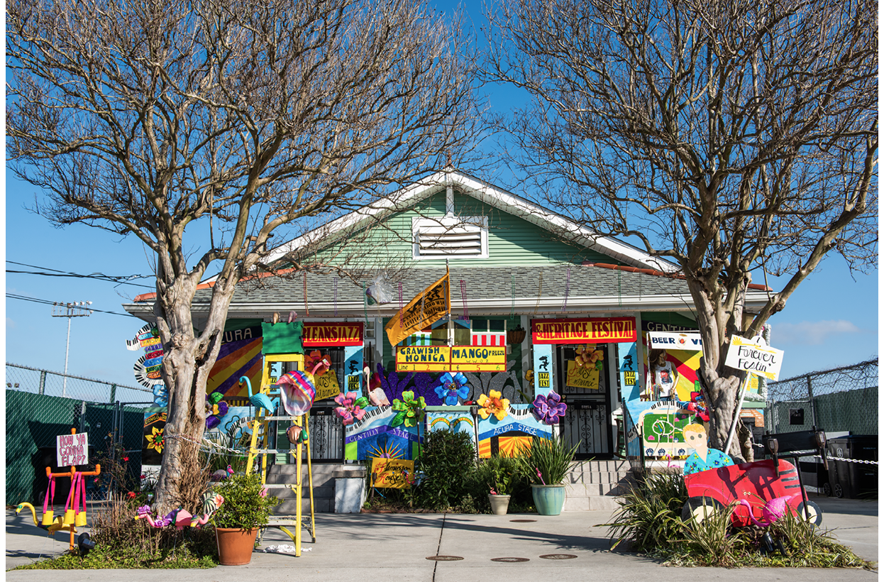 A one-storey house on Fortin Street, New Orleans, is decorated with many signs that borrow from the iconography of the New Orleans Jazz & Heritage Festival, including piano keyboards, a map of the Festival site, names of the stages (‘Acura Stage’, ‘Gentilly Stage’) and classic festival food (‘Crawfish Monica’, ‘Mango Freeze’). A cut-out of a man driving a tractor attached to a tree in front right of the house represents Quint Davis, the producer of the Festival. He holds a sign with the title of the house float, ‘Forever Festin’’. The high fence behind the house is the fence around the Fair Grounds Race Course, where the Festival is held. The photo is taken on a sunny day and the sky is bright blue, which contrasts well with the yellow and red in many of the signs.