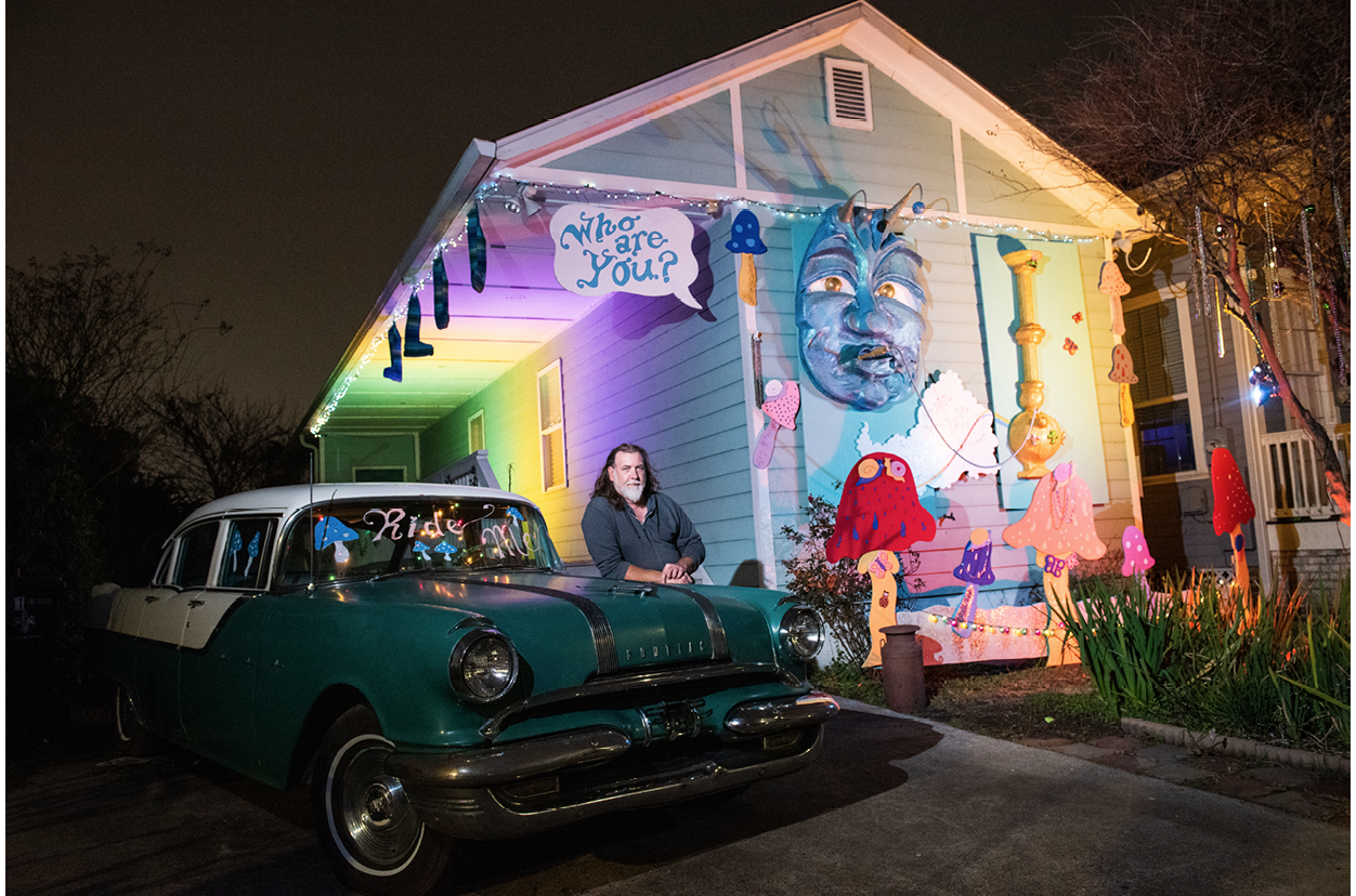 Thom Karamus, a middle-aged bearded white man with long hair, leans on a classic car outside his one-storey house on Dryades Street, New Orleans, which is decorated as the Caterpillar from Alice in Wonderland. A large paper-mâché relief of the Caterpillar’s face is hanging on the short end of the house, along with a hookah that the Caterpillar is smoking and several spotted mushrooms. Caterpillar legs dangle off the porch roof on the long side of the house. A speech bubble sign saying ‘Who are you?’ also hangs on the house. The photo is taken at night and the porch is lit with green, yellow, and purple lights (Mardi Gras colours).