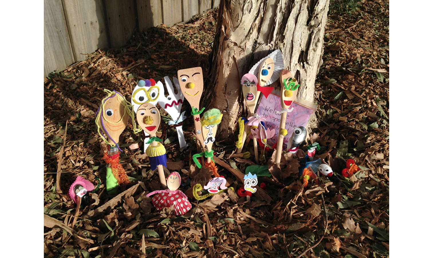 Photo of a  group of wooden spoons standing upright in the ground in the bark at the foot of a tree, and decorated with colourful craft materials to look like people.