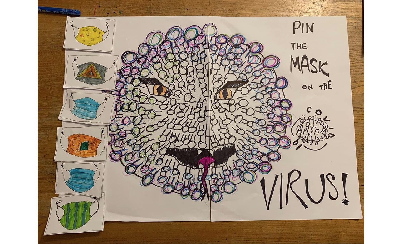 Photo of children's colourful drawings, a large one of a giant Covid-19 virus with a scary face, with the words Pin the Mask on the Virus beside it, and smaller ones of different coloured face masks to pin on it for a blindfold party game.