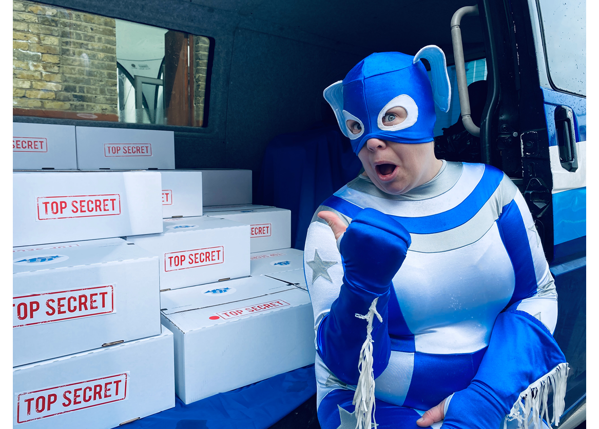Touretteshero, the world’s first Touretteshero superhero, is sitting in the Touretteshero van pointing excitedly at a large set of top-secret superhero kits. Touretteshero is wearing her white and blue superhero costume and mask with flappy ears.