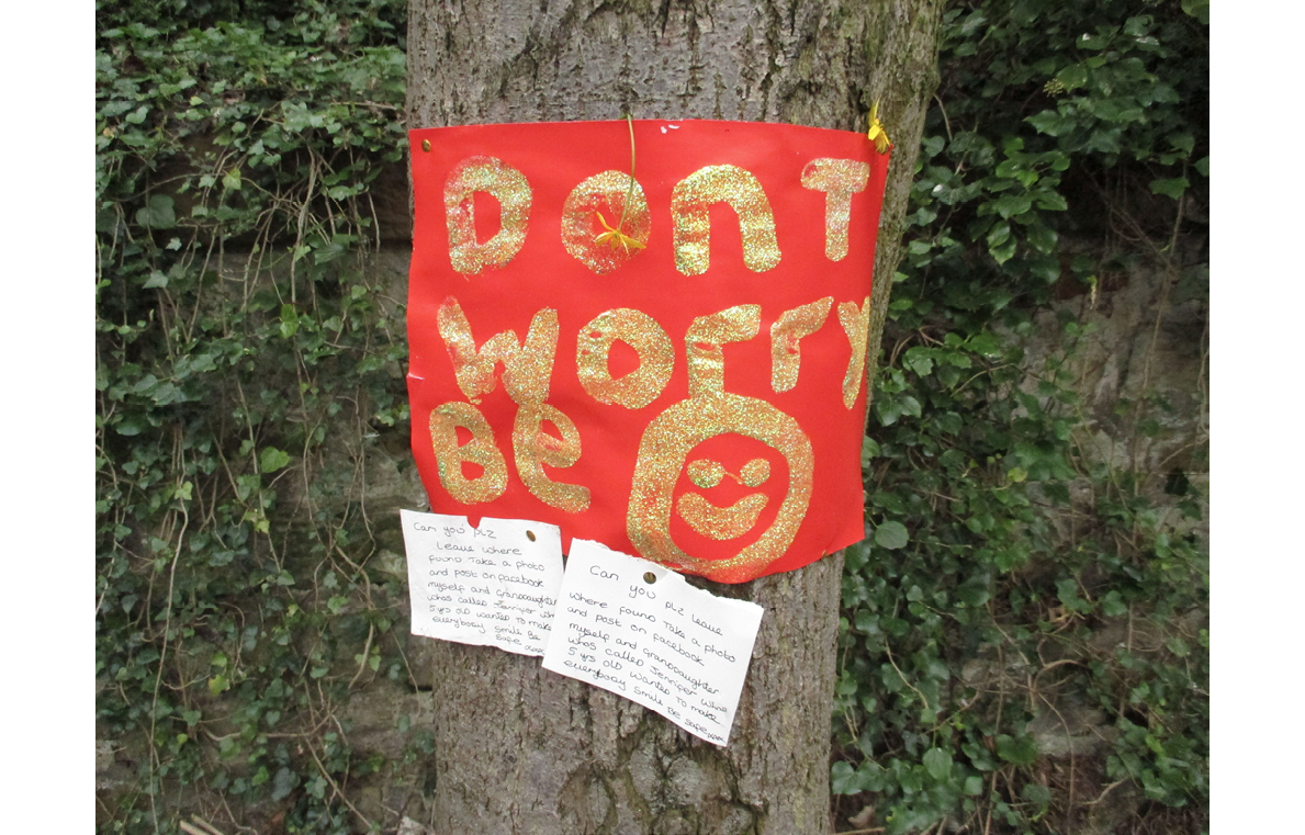 Photo of a home-made notice on orange paper, attached to a tree, with message 'Don’t worry, be' plus a smiling face drawing, in gold lettering, with two handwritten notes on white paper beneath