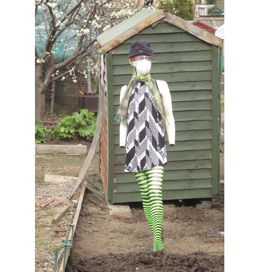 Mannequin placed in front of a green-painted shed, wearing a black-and-white print mini-dress, green and black striped tights, and a woolly hat and scarf, goggles and medical face mask.