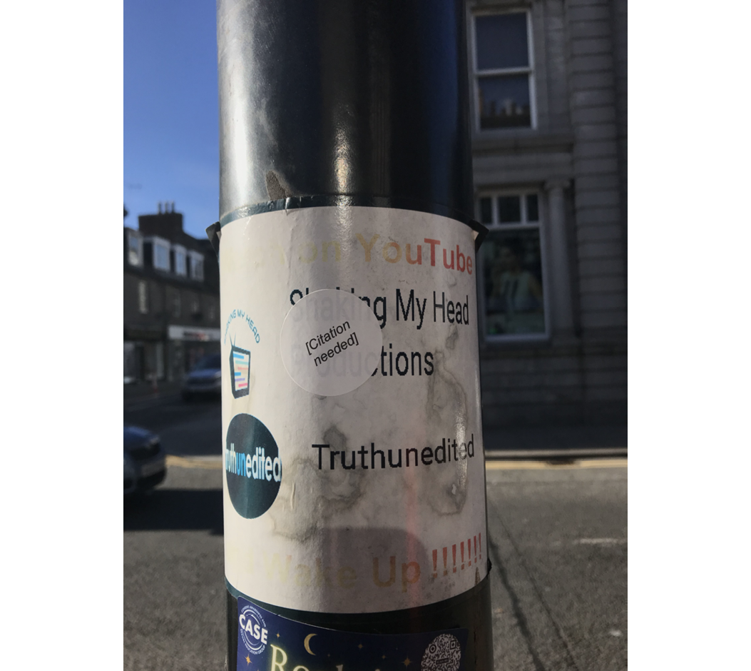Close up photo of a notice attached to a lamp-post, promoting websites relating to discussion of 'fake news', onto which a sticker reading 'citation needed' in square brackets has been added