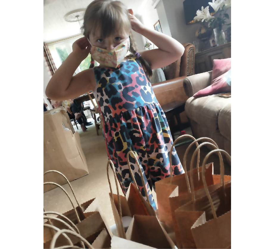 Photo of young girl in a living room, trying on a fabric face mask, with brown paper party bags on a surface in front of her