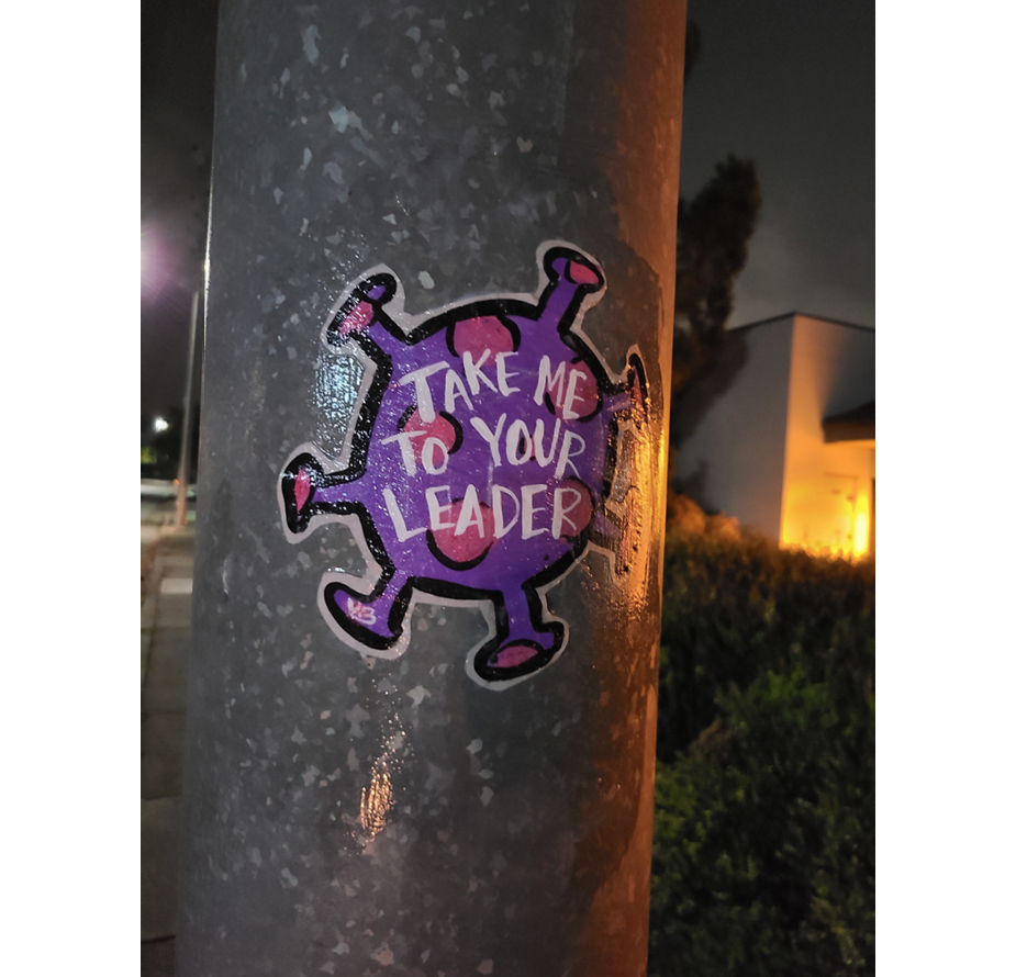 An image adhered with wheat paste. The Coronavirus is depicted as a spiky blue and turquoise sphere, reminiscent of a bouncy ball. A bright pink label on the sphere reads 'Take Me To Your Leader.'