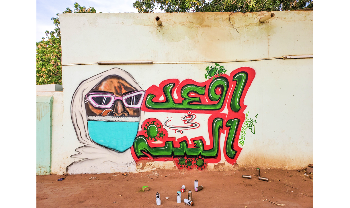 A painting in spray paint on a wall depicts a man in a white hoodie with pink sunglasses. He wears a surgical face mask. Text in Arabic reads 'Stay At Home' and small renditions of the spikey coronavirus surround the text.