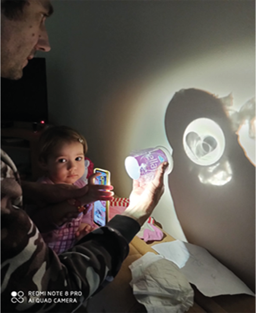 Photograph of father and child shining mobile phone torch through disposable cup and lid, creating projection on wall