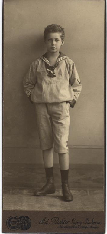 A sepia-toned photograph of a young schoolboy wearing a white sailor outfit with a dark striped tie around his neck and black boots