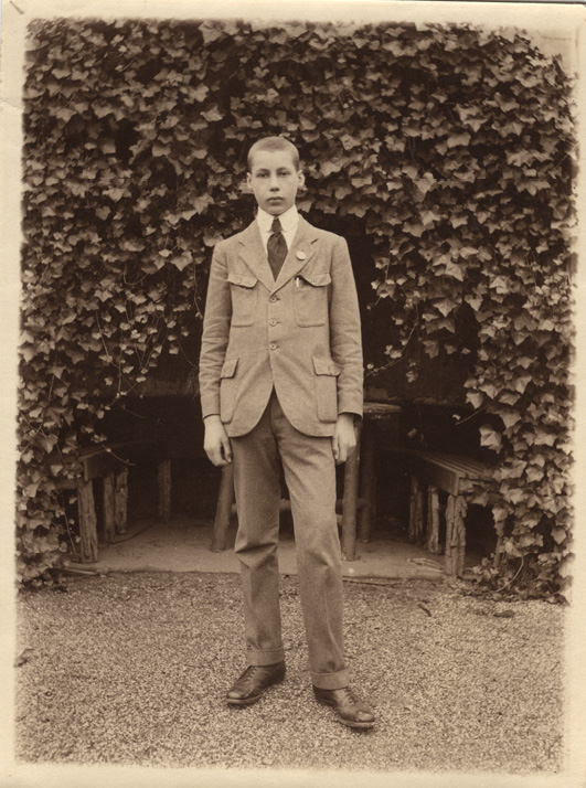 A sepia-toned photograph of a young light-haired teenage boy wearing a light-coloured suit and dark tie standing by an ivy-covered wall outside