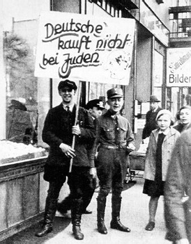A black and white photograph of a young man in a dark suit holding a large hand-written sign that says ‘Boycott of Jewish Businesses’ standing in front of a shop next to a young man in a dark uniform and hat