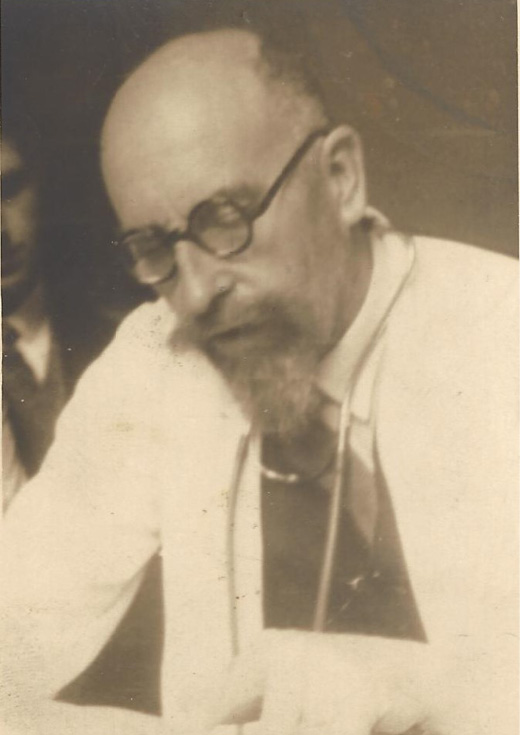 A sepia-toned photograph of a bald man with a grey beard and moustache wearing black round spectacles, a lab coat, white gloves and a stethascope around his neck