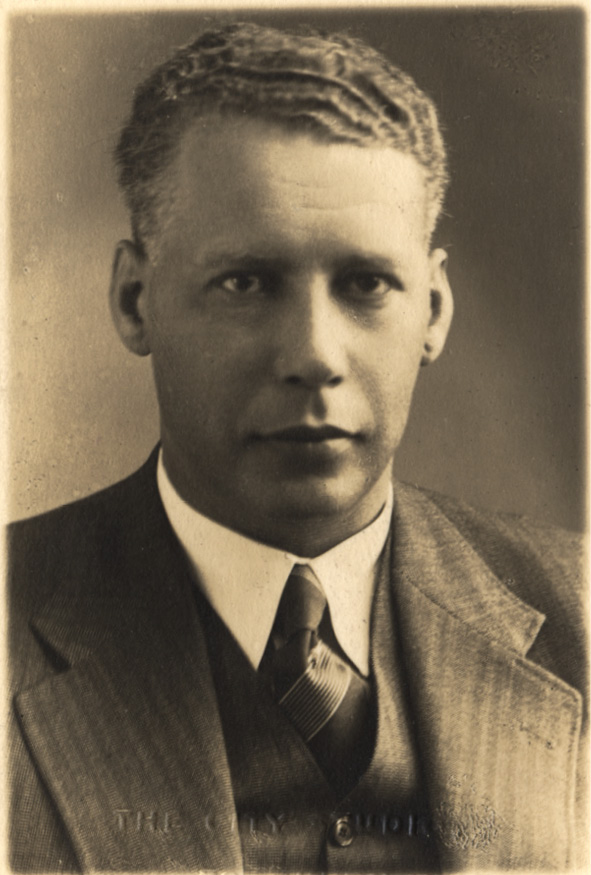 A sepia-toned photograph of a light-haired man in his forties wearing a pinstripe double-breasted suit, a white shirt and a striped tie