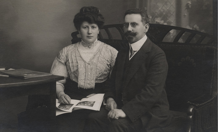 A black and white photograph of a woman with dark hair in an up-do wearing a light blouse holding a book with photographs sitting next to a dark-haired man with a moustache in a dark suit