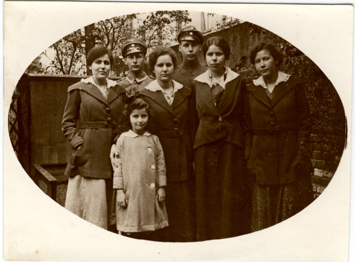 A sepia-toned group photograph of a young dark-haired girl in a button-up coat standing with four women in dark coats and white shirts in front of two men in military uniforms