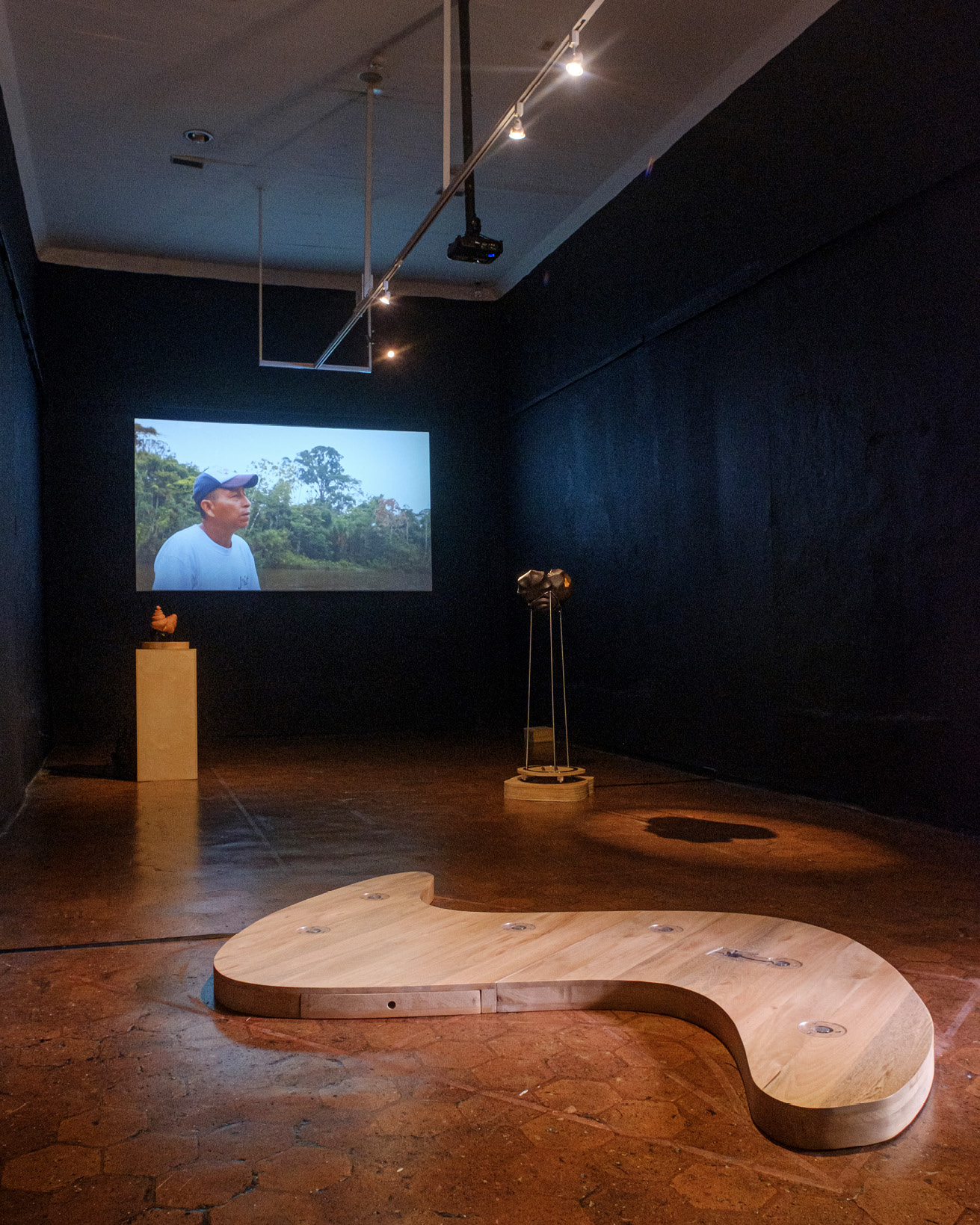 Dimply lit exhibition room, with two floor mounted artifacts and a video caption projected on a wall showing a human person speaking.