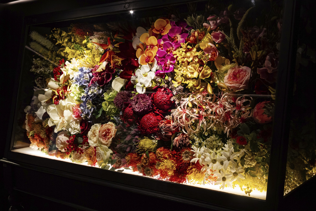Glass display case filled with artificial flowers looking bright in strong light against a dark background.