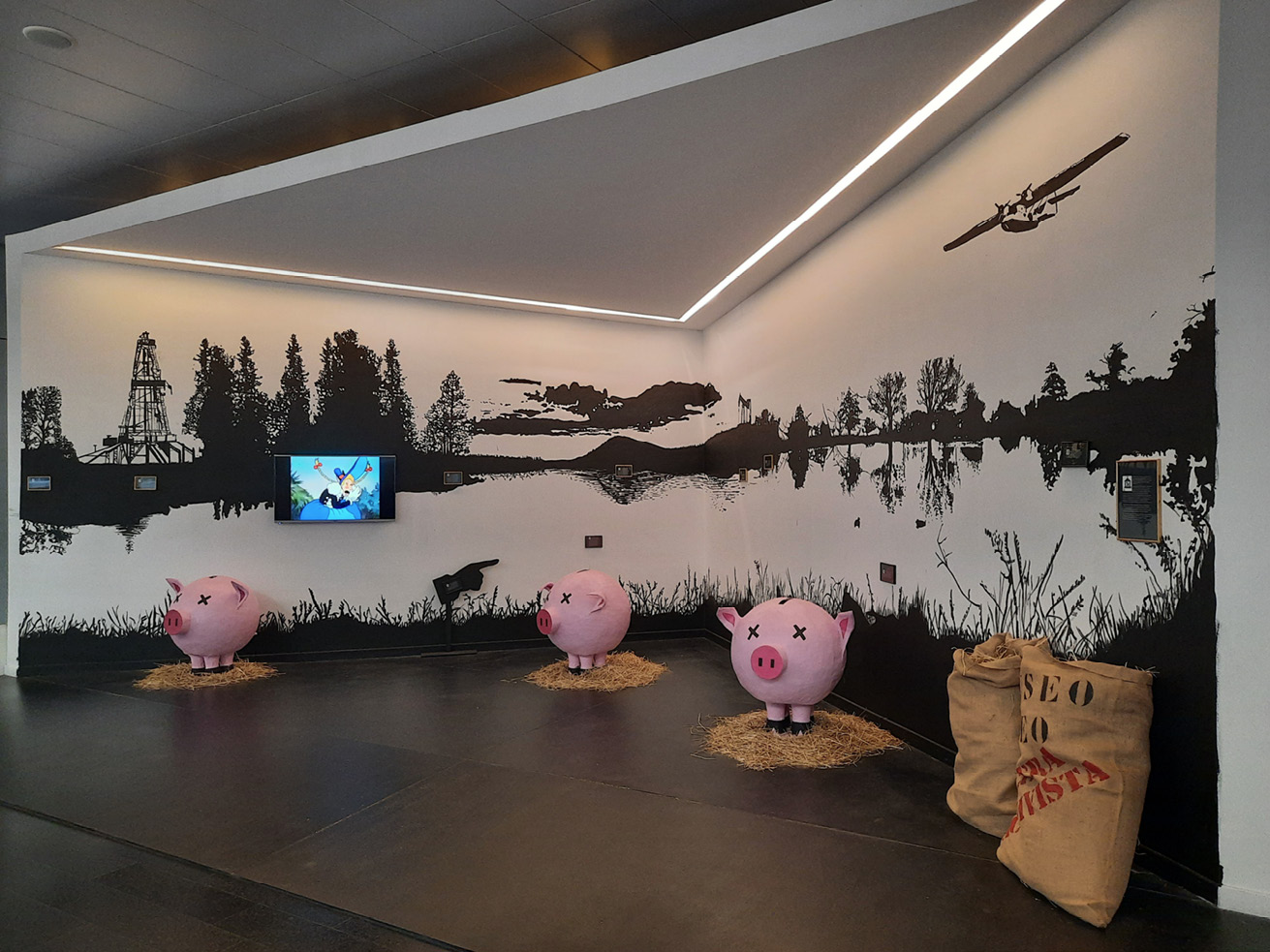 View of a diorama with three pink piggybanks sitting on small hay piles, two sacks with hay, a monochromatic industrial background painted on the walls, and a wall-mounted screen projecting cartoons.