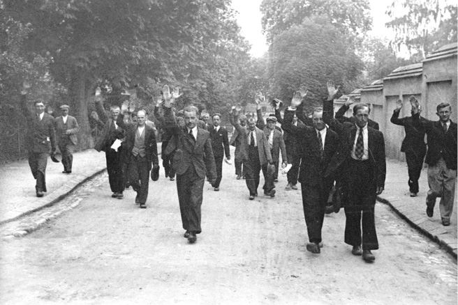 The Mishlenitse pogromists on the march. About two dozen men, most wearing suits and ties, march down a street as they enter Mishlenitse. Their right arms are raised in a Nazi salute.