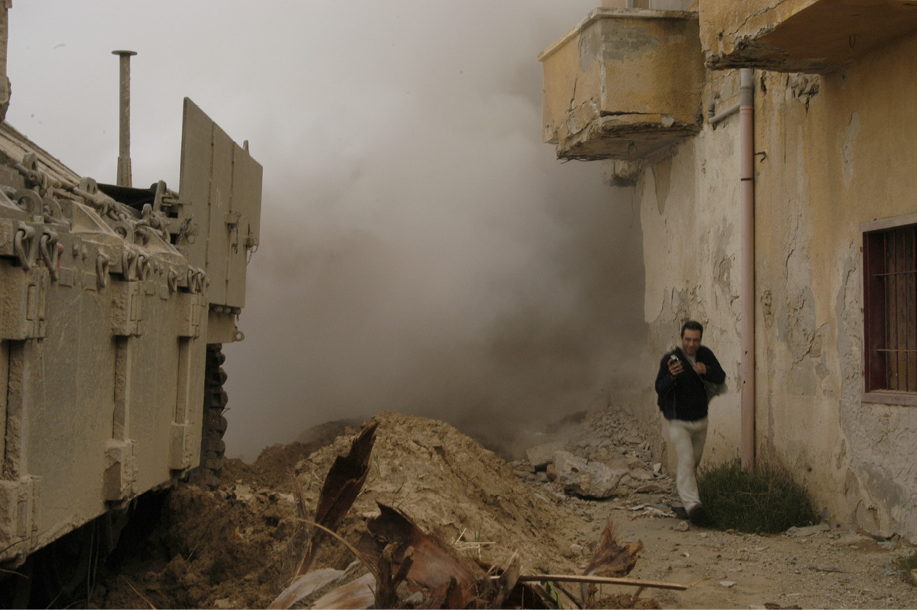 Man walking past a shelled building and a tank, with heavy smoke rising behind him.