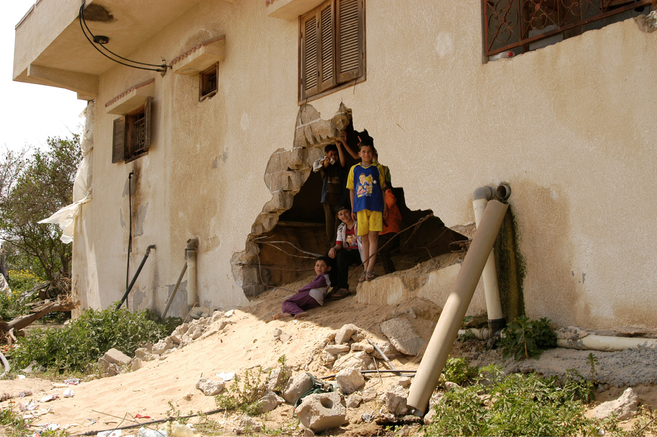 Six Palestinian children standing or sitting down in the blown hole of a shelled house, some looking straight into the camera while one of them is making the victory sign.