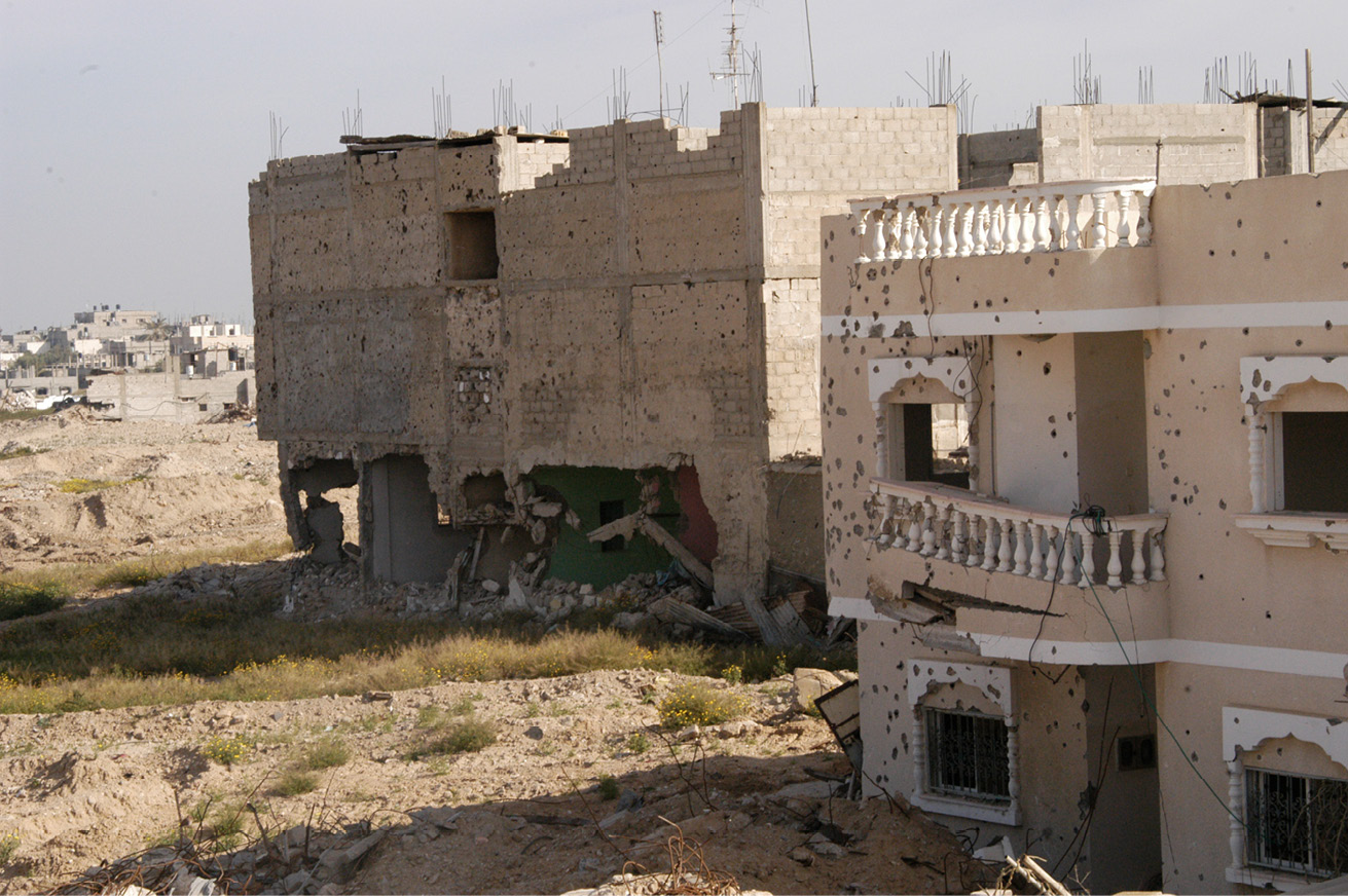 Close-up of two heavily shelled residential buildings, with bullet holes on walls and metal frames exposed.