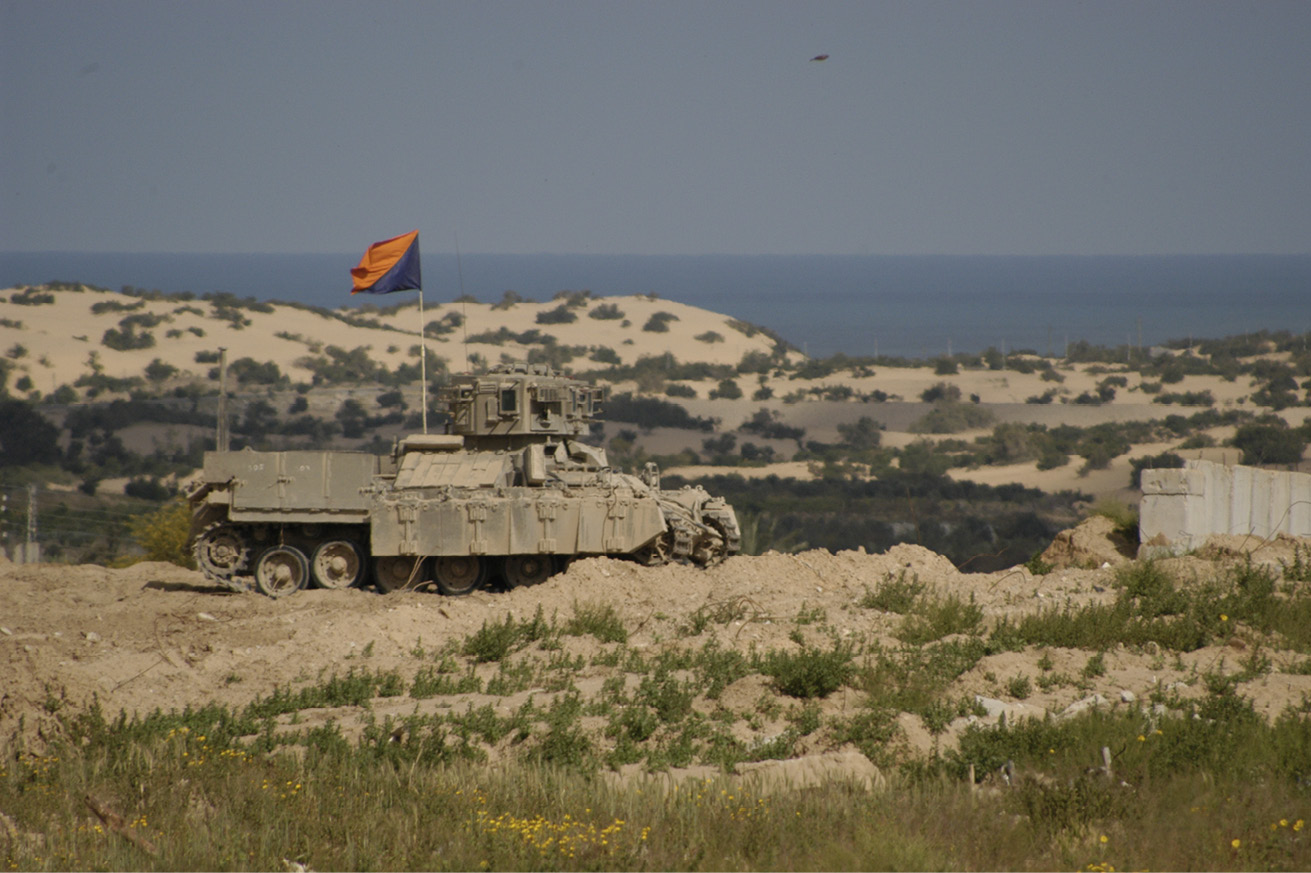 Tank hoisting a diagonally divided blue and orange flag and positioned near a body of water.