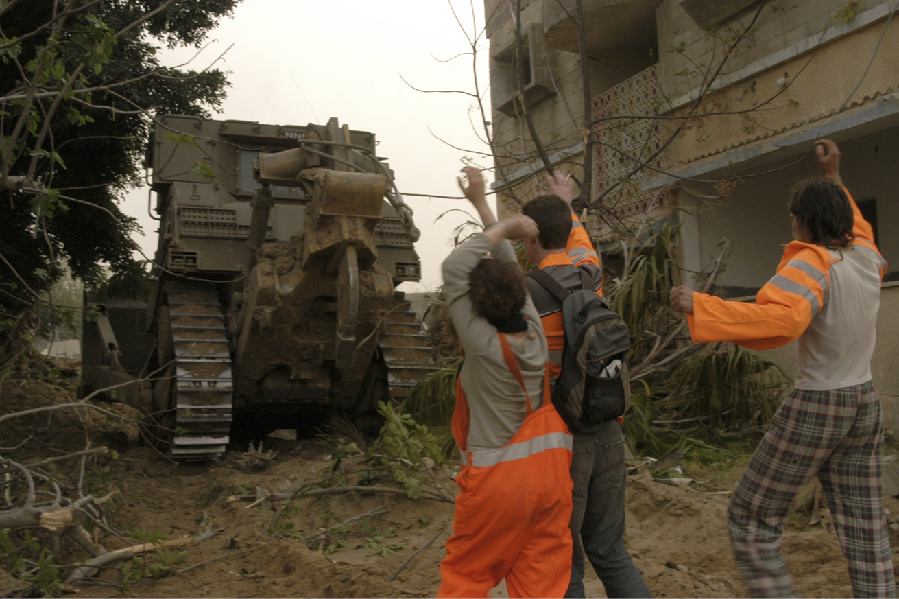 Three persons wearing orange signal clothes standing in front of a tank and gesturing towards it, with their hands in the air.
