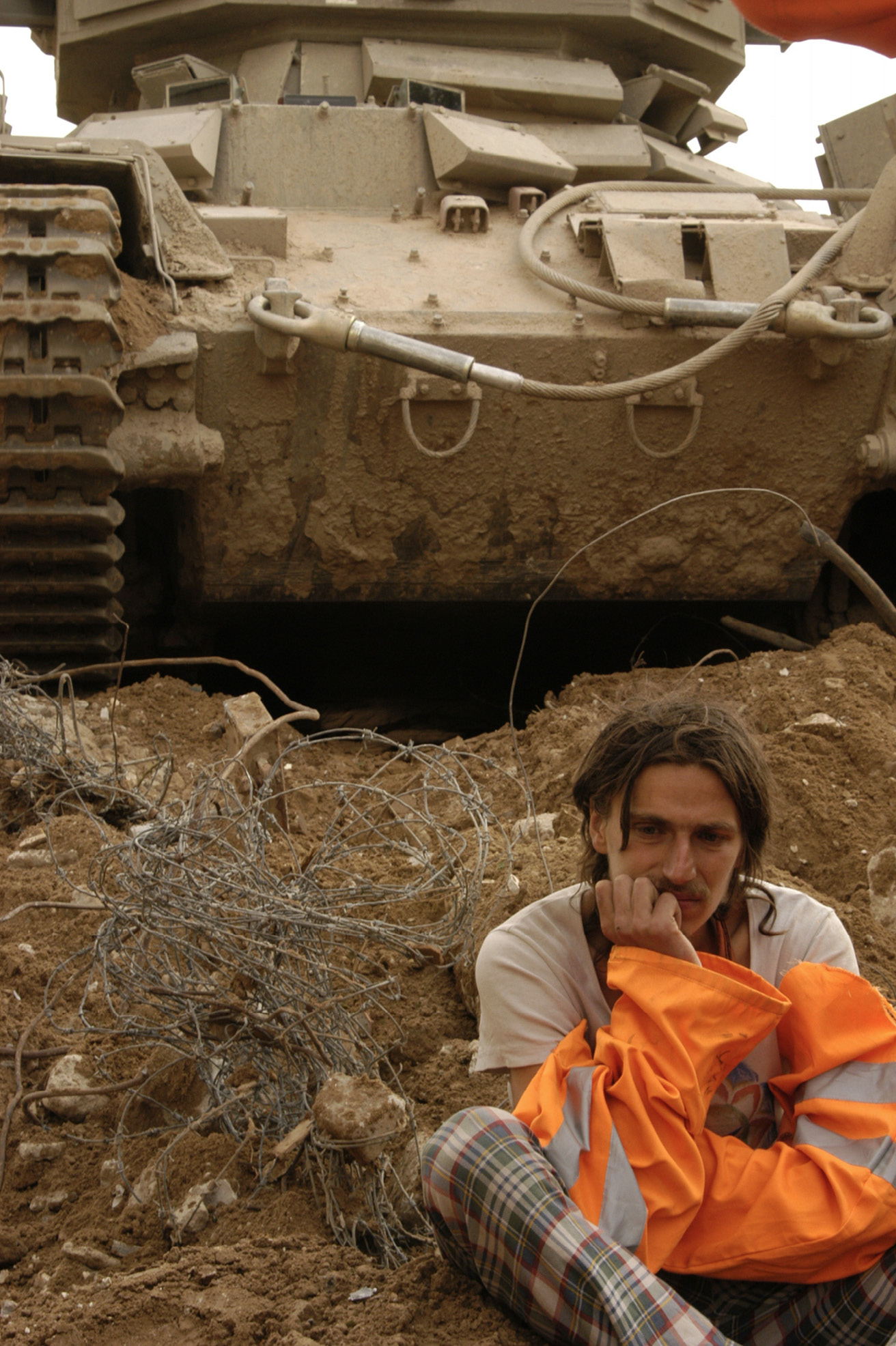 Frontal close-up of a man in orange signal clothes holding face on one hand while sitting down near a barbed wire clew and in front of a tank.