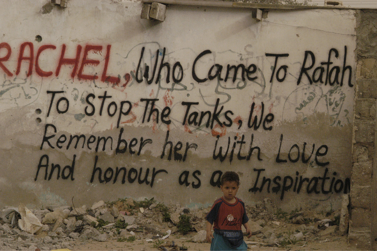 Toddler standing in front of a wall with a graffiti reading ‘Rachel, who came to Rafah to stop the tanks, we remember her with love and honour as an inspiration’.