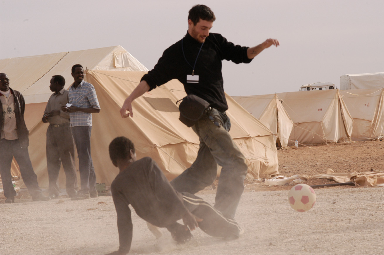 Man playing football with a child, with three men standing at a distance in front of raised tents.