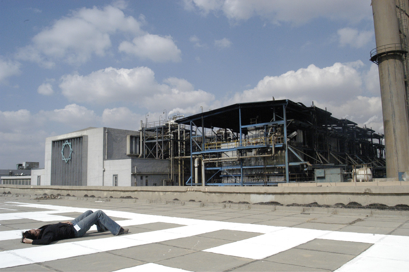 A person lying flat on their back on the roof of a power station surrounded by other industrial buildings with pipes and tubes outside, one building also displaying the symbol of an eight-pointed blue star.