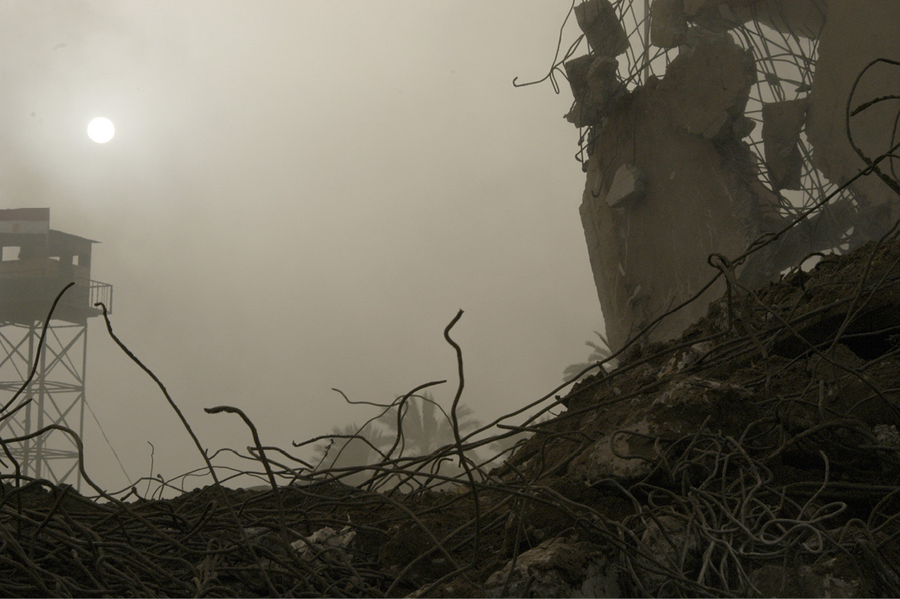 Rubble debris and tangled metal wire, with the moon and an observation tower in the background, both seen through heavy smoke.