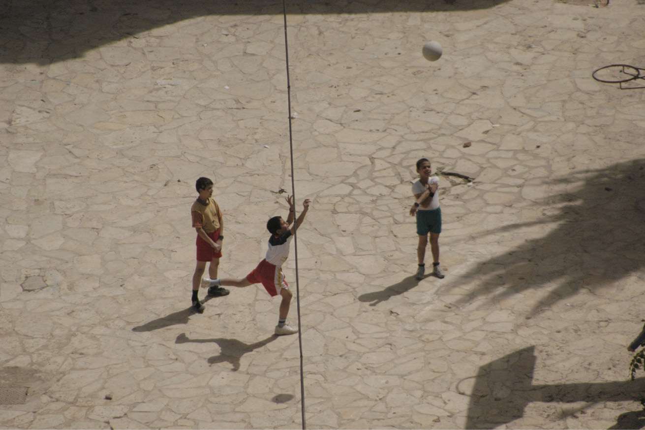 Three Palestinian children playing basketball in the street, two of them standing and watching while the other one is throwing the ball to the basket.