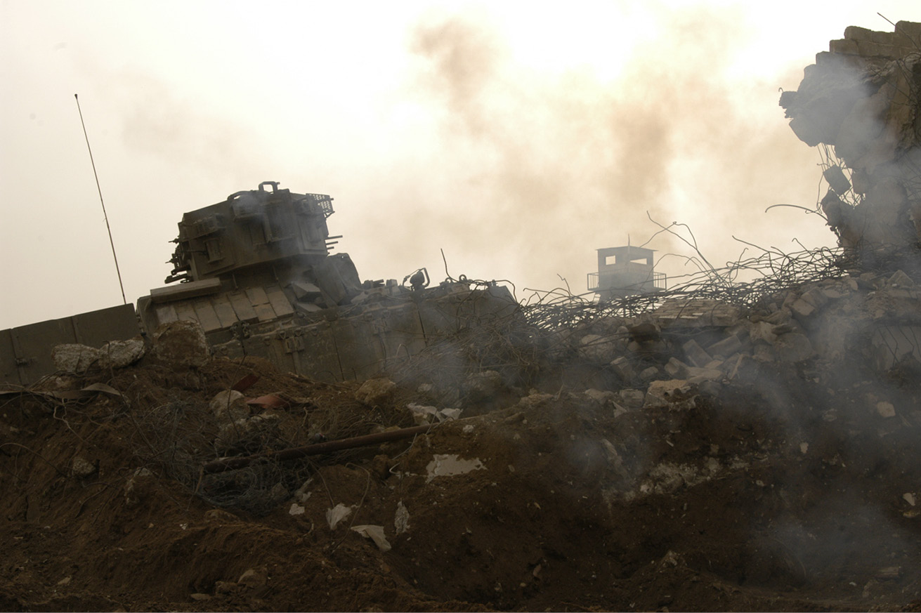 Pile of rubble seen through smoke, with a tank and an armoured observation post in the background.