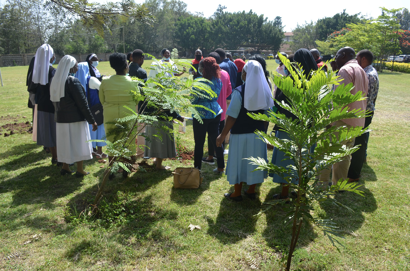 Group of people of colour, some of them wearing face masks or female religious clothing, all gathered around a newly planted tree and listening to a person speaking.